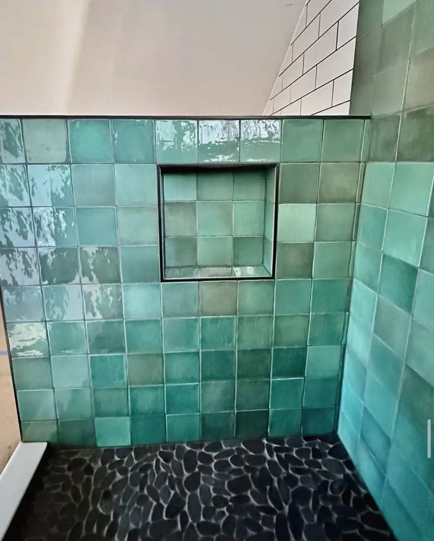 Green ceramic tile shower wall with built-in niche and black pebble floor