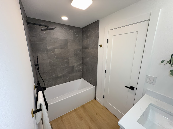 Finished bathroom with gray marble-look tub surround, matte black fixtures and wood-look flooring